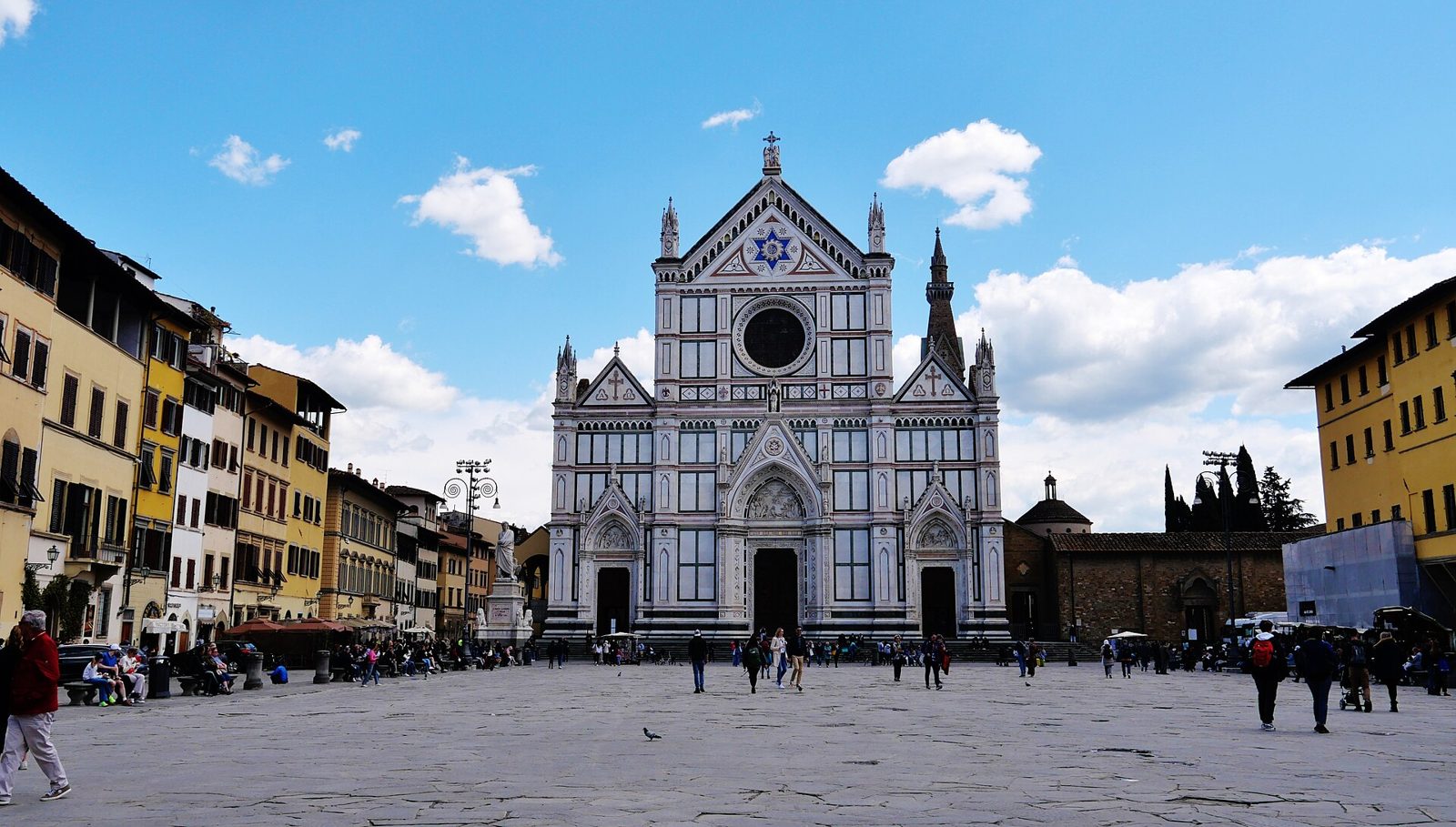 Piazza Santa Croce in una giornata di sole: la facciata in marmo bianco e verde della basilica con il campanile, il grande selciato quasi libero con qualche passante lontano e i palazzi colorati ai lati