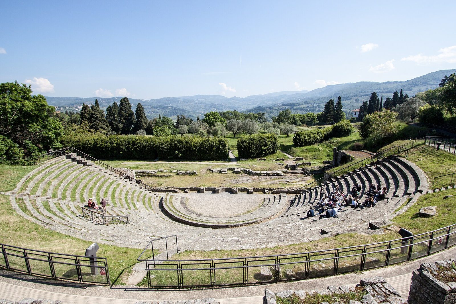 Le gradinate semicircolari del teatro romano di Fiesole viste dall'alto, con l'orchestra al centro, i resti del palcoscenico e sullo sfondo cipressi, ulivi e colline sotto un cielo azzurro