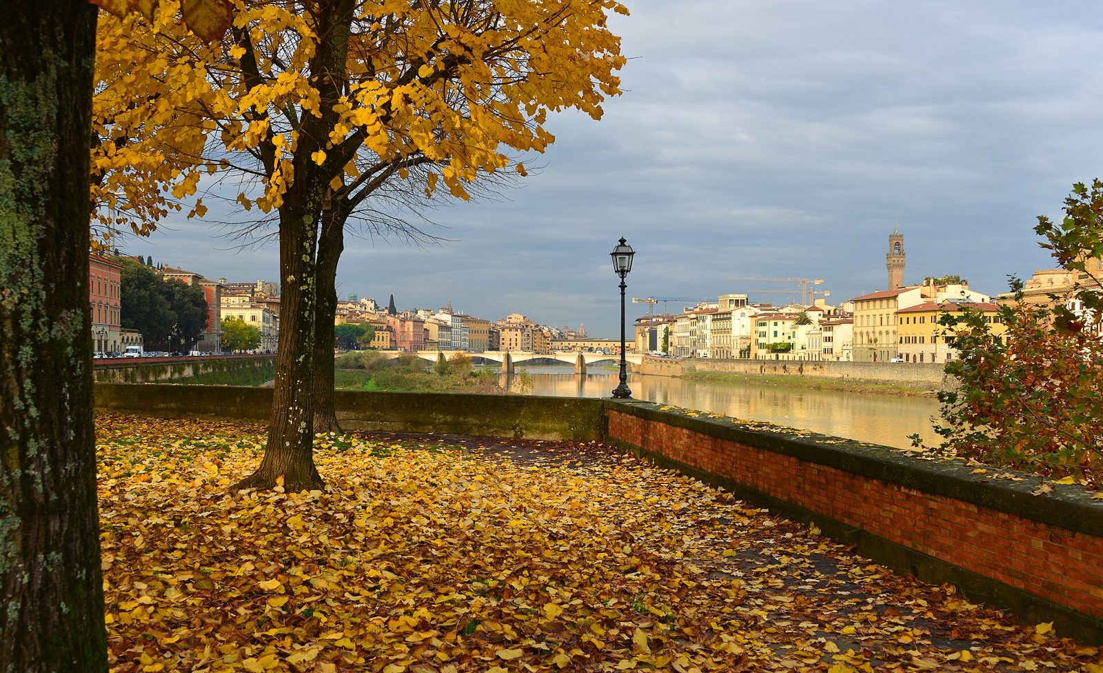 Lungarno in autunno con foglie gialle sul selciato e sugli alberi, l'Arno, un ponte e la torre di Palazzo Vecchio sotto un cielo grigio