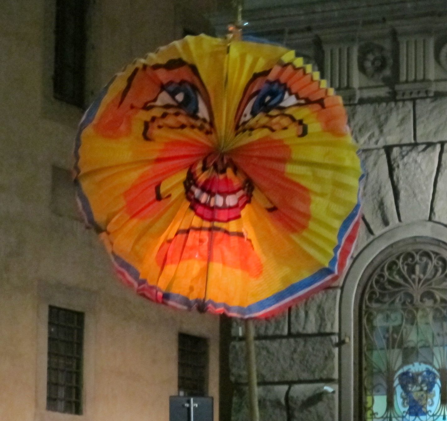 Una rificolona di carta a fisarmonica, gialla e arancione con un volto di sole dipinto, illuminata dall'interno e sollevata su un'asta davanti alla facciata in pietra di un palazzo, di sera